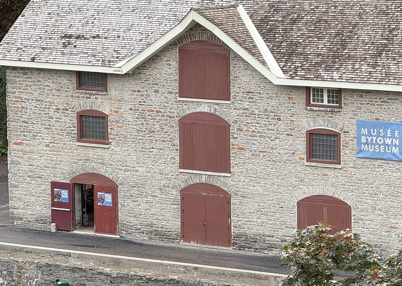 The grey limestone facade of the Commissariat Building, now the Bytown Museum, its red-painted supply doors stacked on three levels, the ground-floor entrance standing open.