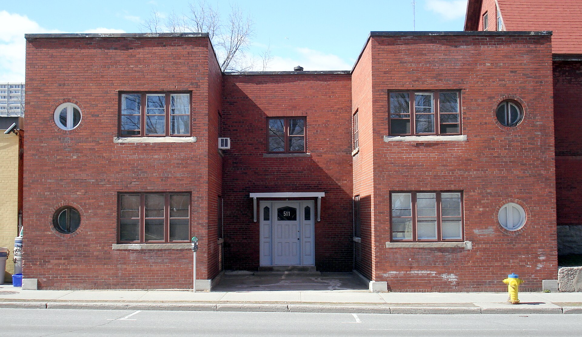 511 Somerset Street West: an ordinary two-storey red brick apartment building with small round windows flanking a recessed entrance, seen straight-on from across the street; the number 511 on the pale front door.